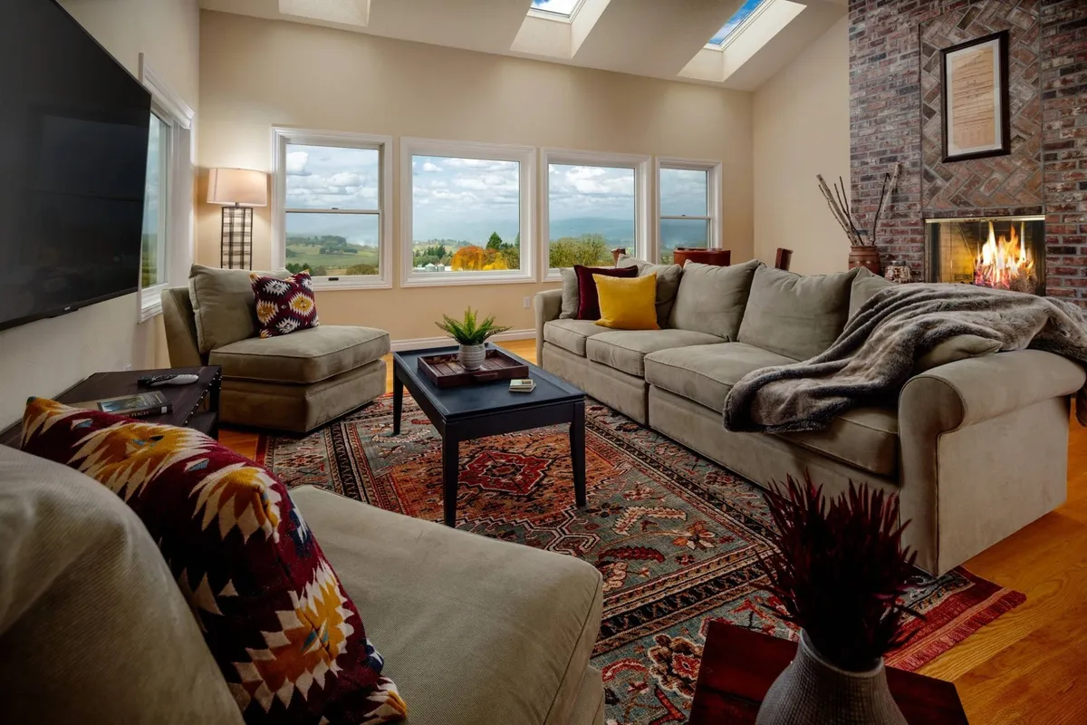 A spacious living room with large windows frames sweeping views of the surrounding landscape. The room is anchored by a warm fireplace, accented with decorative throw pillows and an inviting area rug. Sunlight pours in, creating a welcoming space ideal for relaxing, entertaining, and enjoying scenic sunsets.