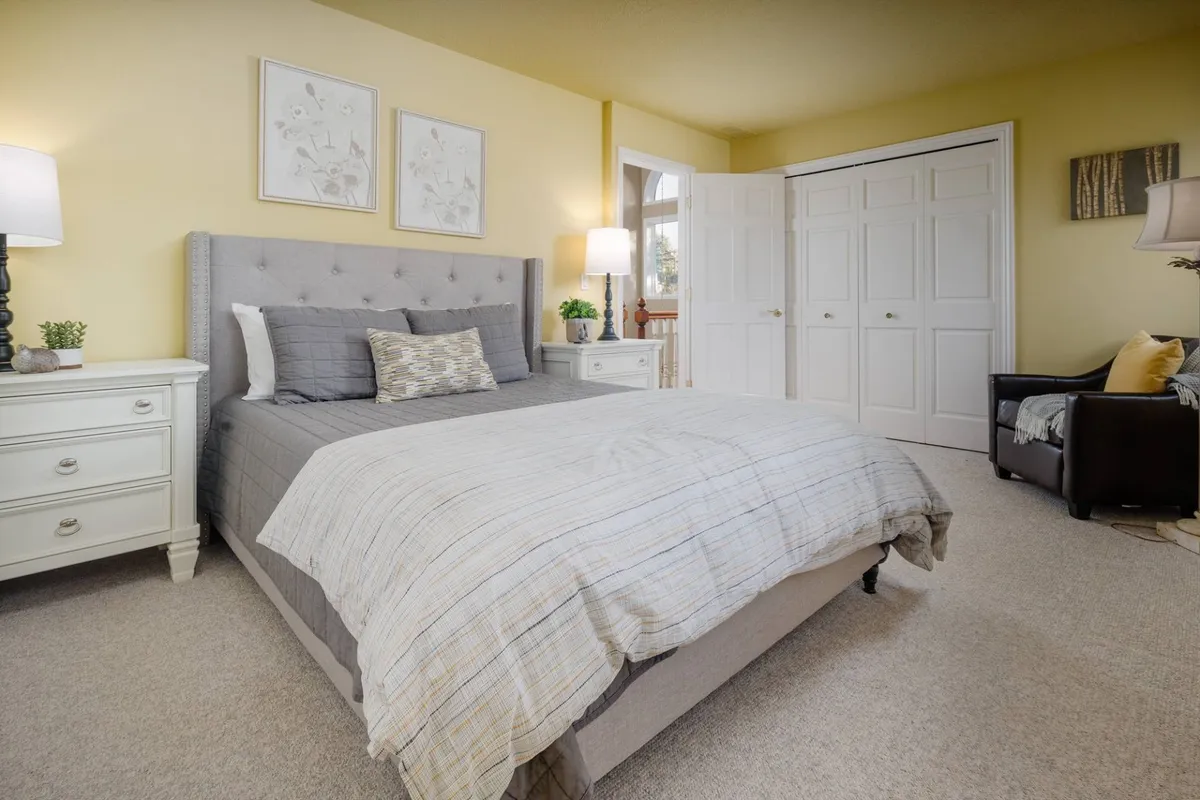 A tranquil bedroom scene with a bed featuring a gray headboard, matching bedside lamps, a white nightstand, framed botanical art, and a window allowing natural light to flood the space. The ambiance is restful and inviting, ideal for a peaceful night's sleep or quiet morning.