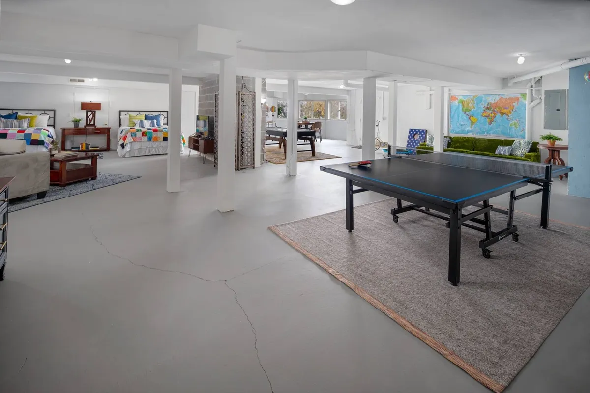 A vibrant basement space featuring a ping pong table, two colorful queen beds, a green sofa, and a wall mural of a world map. The room is lively and welcoming, with plenty of space for both games and relaxation.