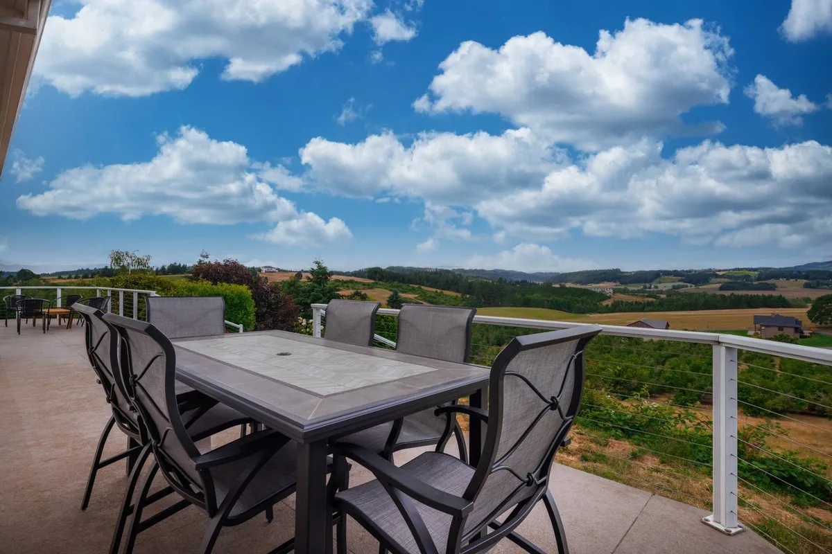 This exterior shot features a large outdoor dining table overlooking a scenic vista under a cloudy sky. The table is set for a meal, with enough space for a large group to gather and enjoy the view. The surrounding landscape reinforces the connection to nature and communal experiences.