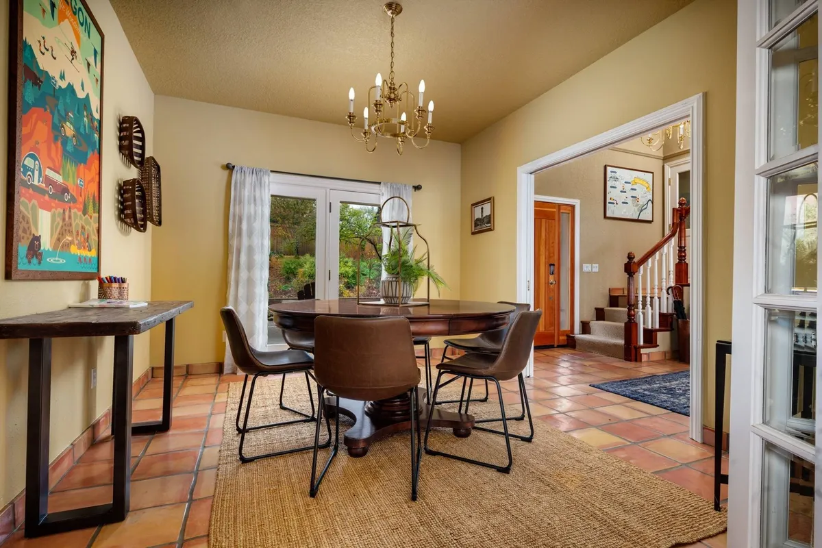 A stylish dining space features a round table under a statement chandelier, with glass doors opening to outdoor scenery. Art adorns the walls, suggesting an atmosphere of creativity and celebration for meals and social events.