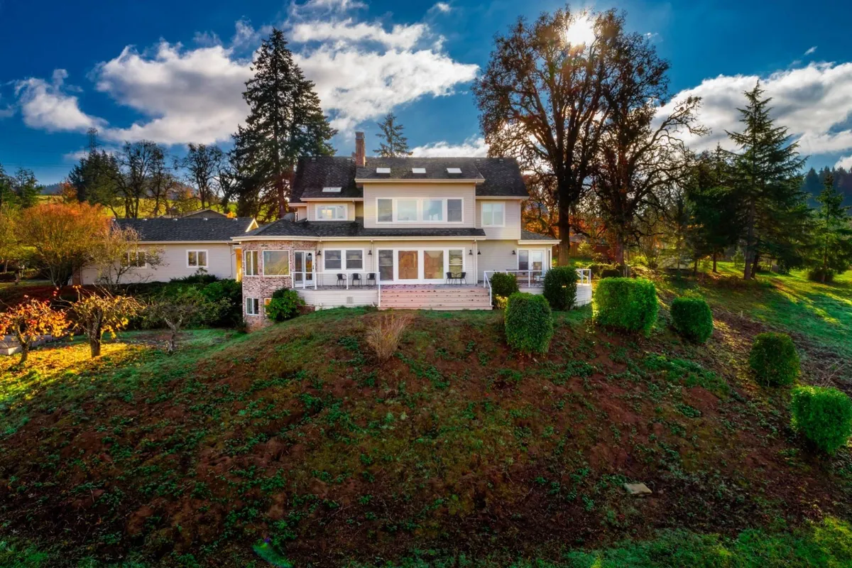 This image shows the main house and porch nestled among lush greenery, with trees and gently rolling hills in the background. The welcoming porch suggests a spot for group gatherings, and the landscape hints at expansive grounds ideal for outdoor games and strolls described in the section.